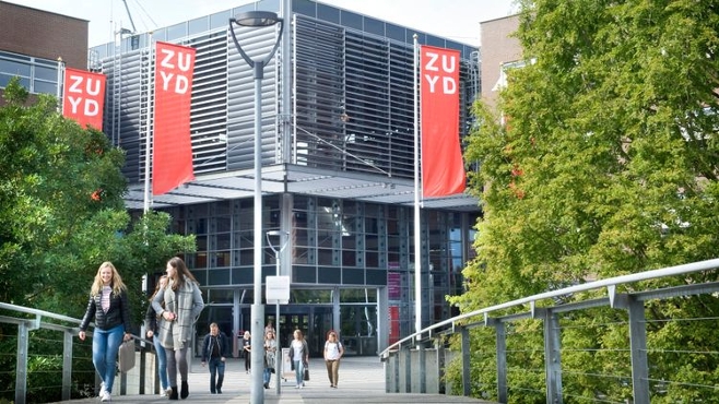 Students walking on a bridge towards the modern glass and steel building of Zuyd University, with red flags
