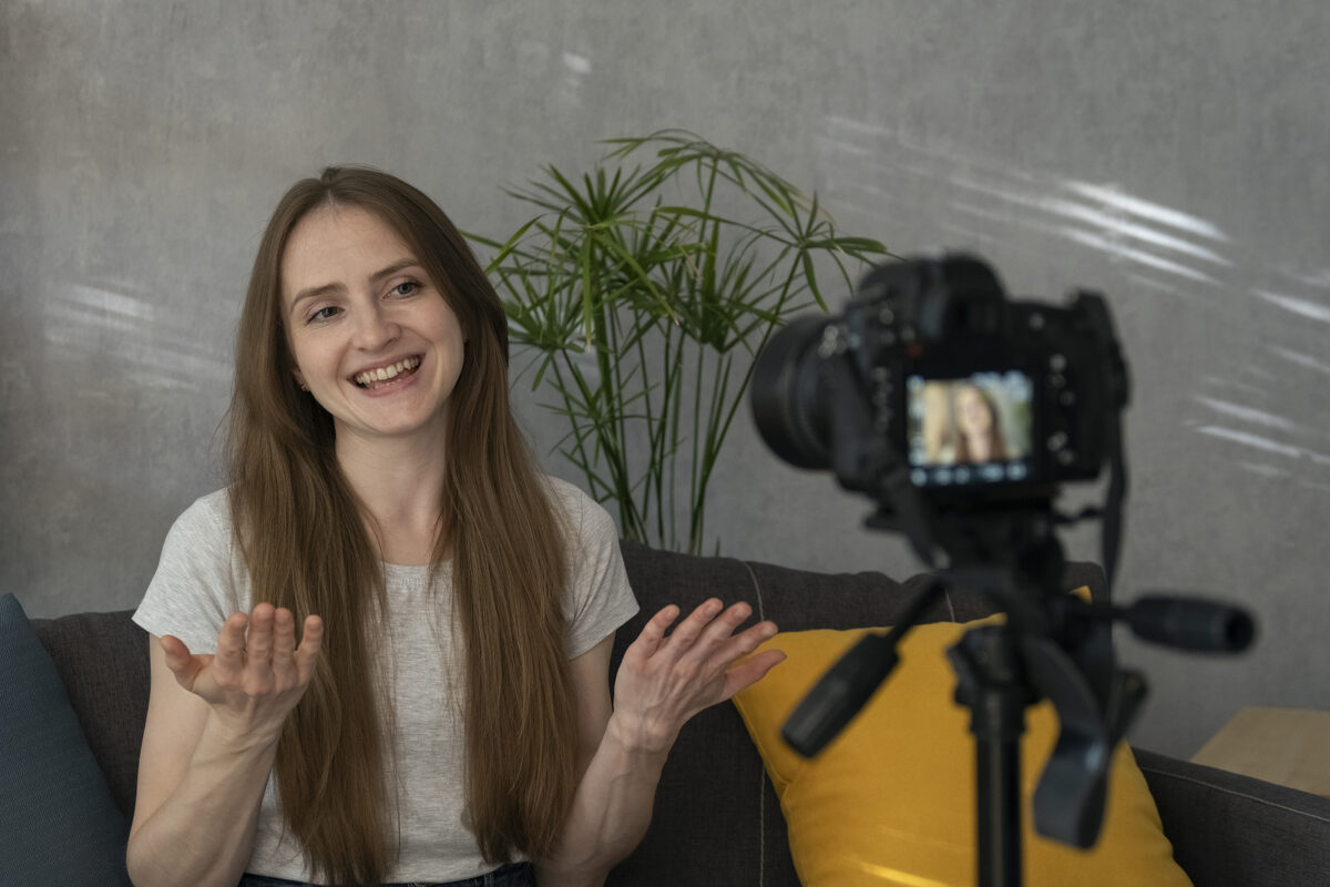 A young woman with long brown hair smiles and gestures while recording a video blog with a camera on a tripod.
