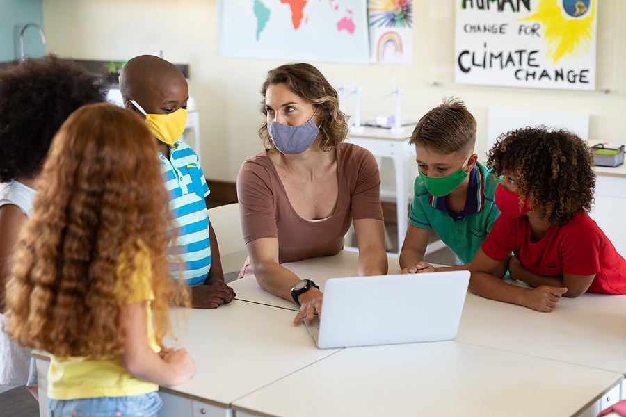 Teacher and four primary school children, all wearing face masks, looking at a laptop in a classroom.