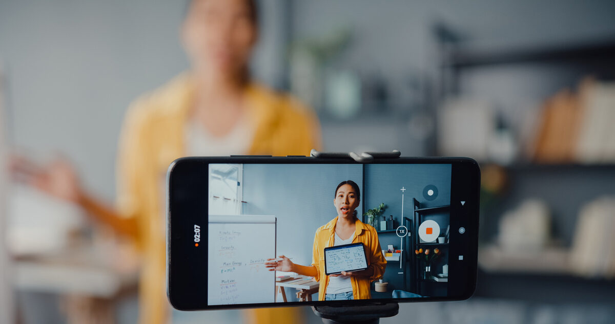 A woman in a yellow shirt records a video of herself presenting with a tablet and whiteboard.