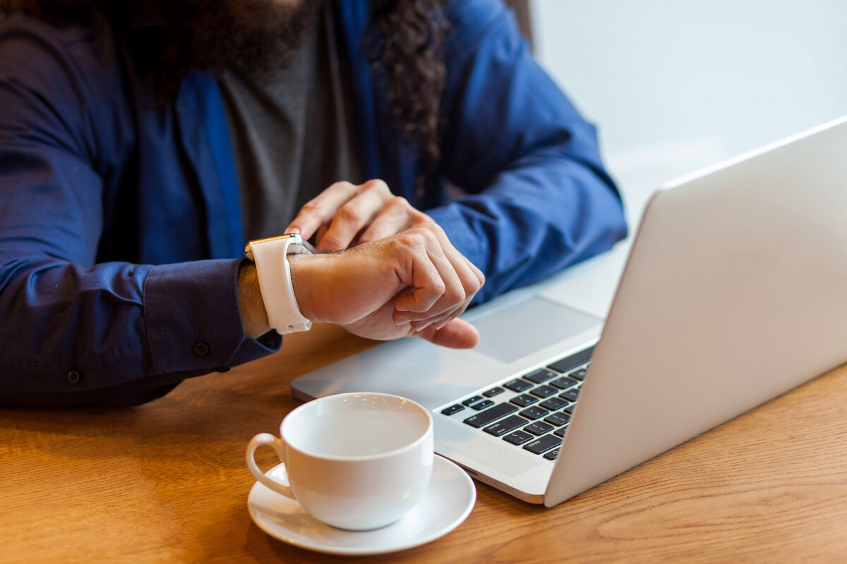 Close-up of a person checking a white smartwatch on their wrist, with a laptop and coffee cup nearby.