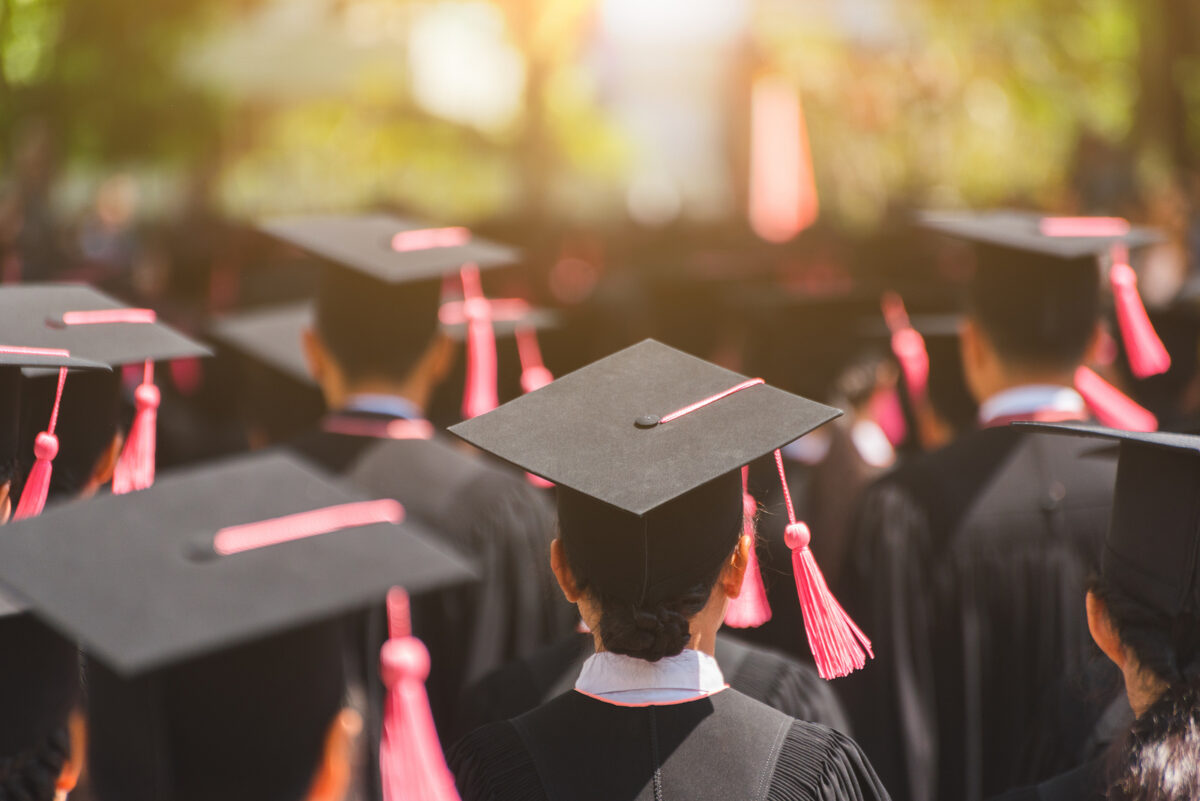 Rear view of graduates in black caps and gowns with pink tassels, facing a bright background