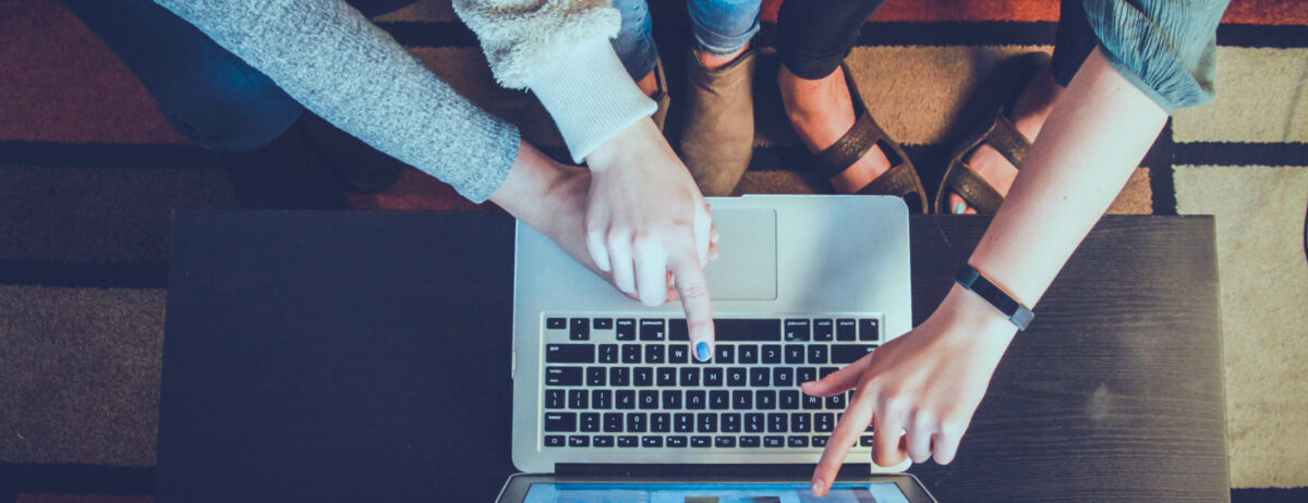 Three people gathered around a laptop, with two hands pointing at the keyboard and screen.