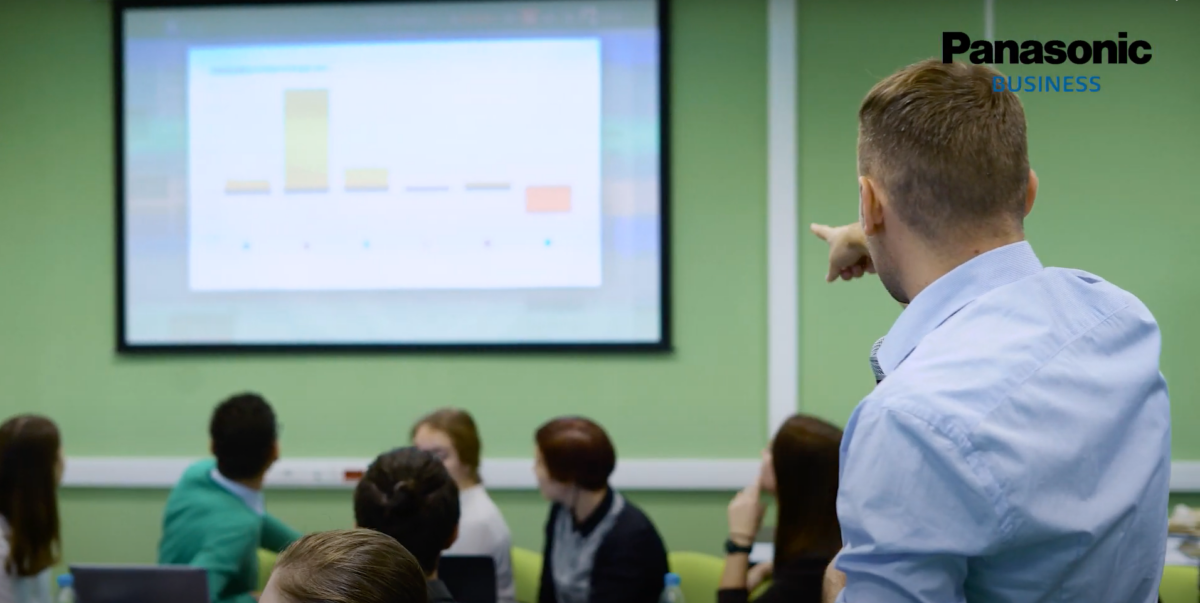 A man in a light blue shirt points to a bar chart on a large screen in a classroom setting.