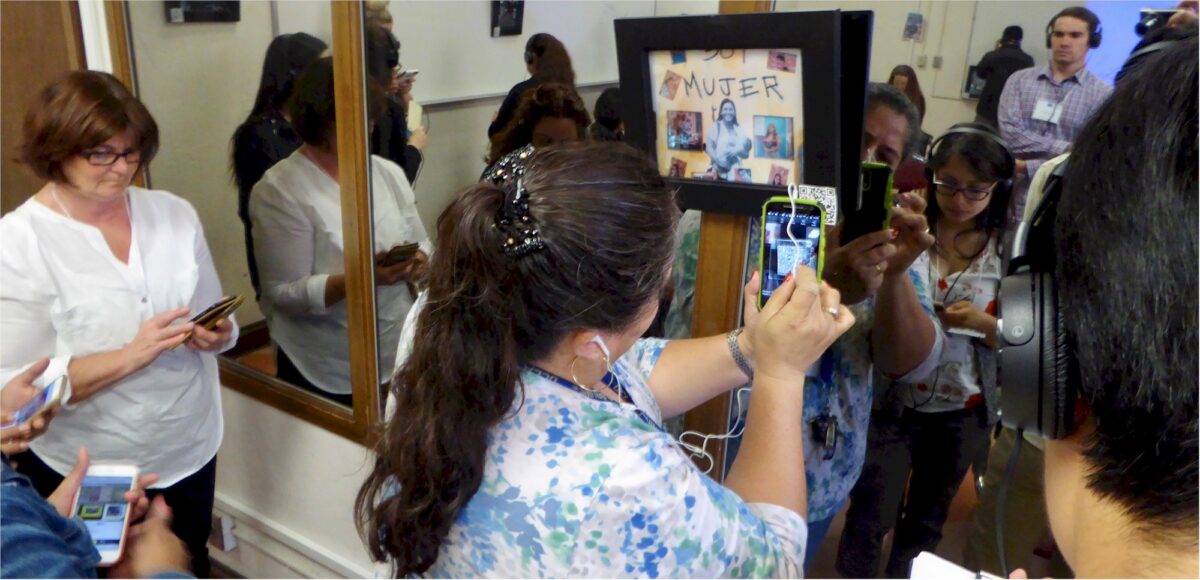 People in a room, some looking at their phones, one person taking a photo of a framed picture.