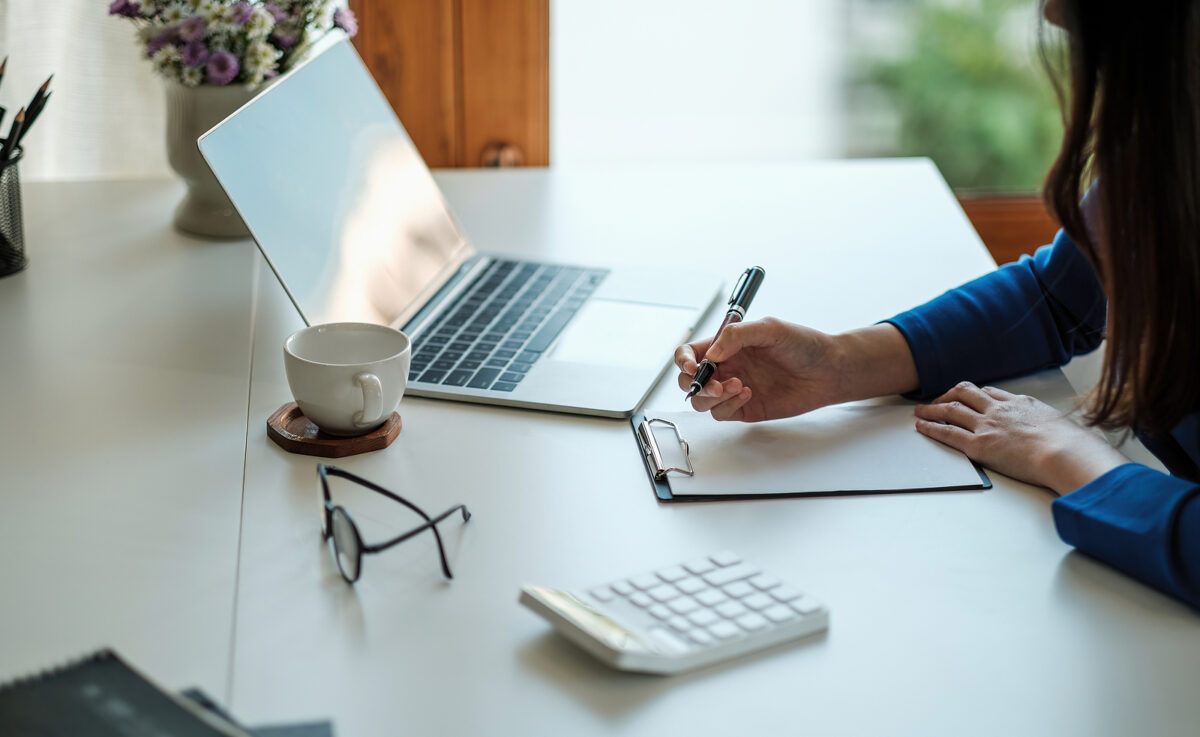 A person writing on a notepad next to a laptop, a cup of coffee, glasses, and a calculator.
