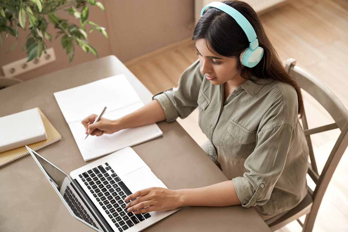 Young woman with headphones writing in a notebook and typing on a laptop at a desk