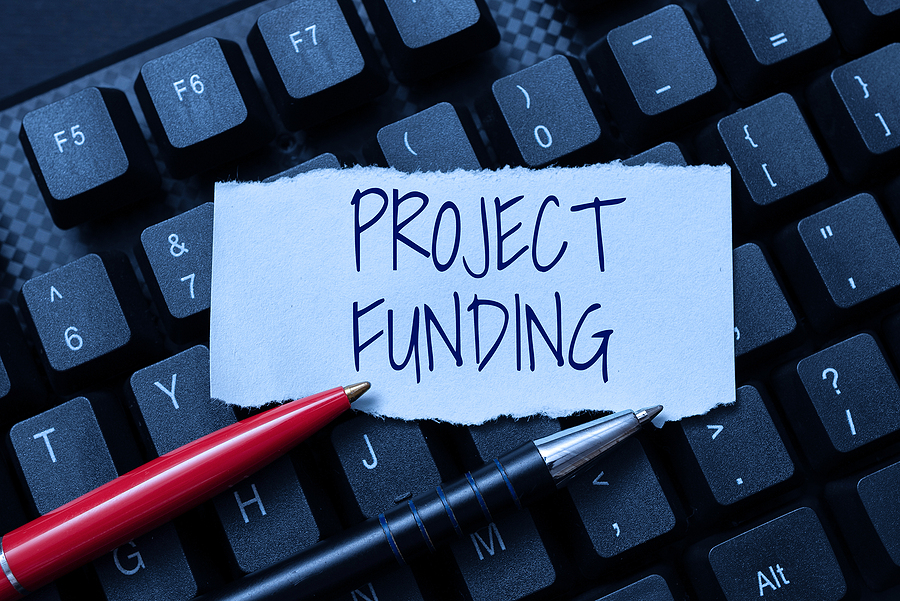 A piece of paper with "PROJECT FUNDING" written on it, placed on a computer keyboard with two pens.