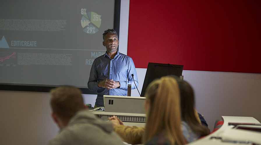 A lecturer in a blue shirt stands in front of a screen with a pie chart and text, addressing students.