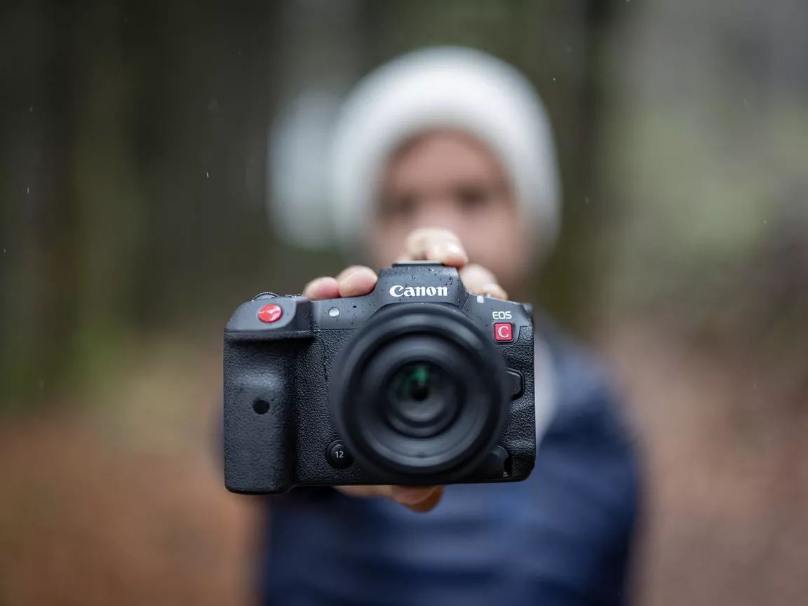 A person holding a black Canon EOS C camera with a lens pointed towards the viewer.