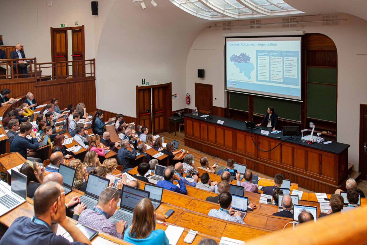 A large lecture hall with many attendees seated at wooden desks, most using laptops, listening to a speaker.