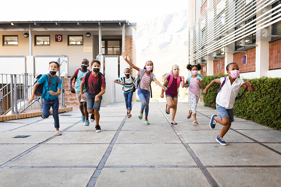 Diverse primary school children wearing face masks and school bags run towards the camera outdoors.