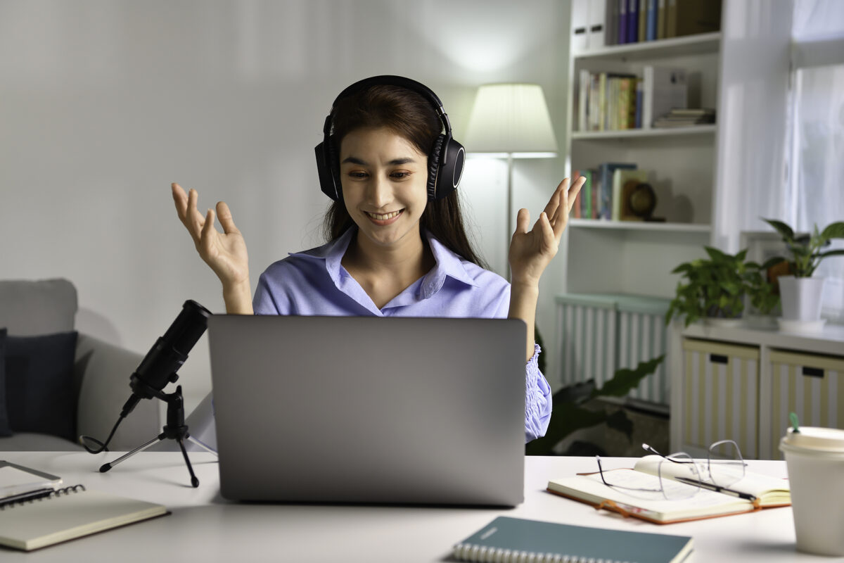 A young woman with headphones on, smiling and gesturing with her hands while looking at a laptop.