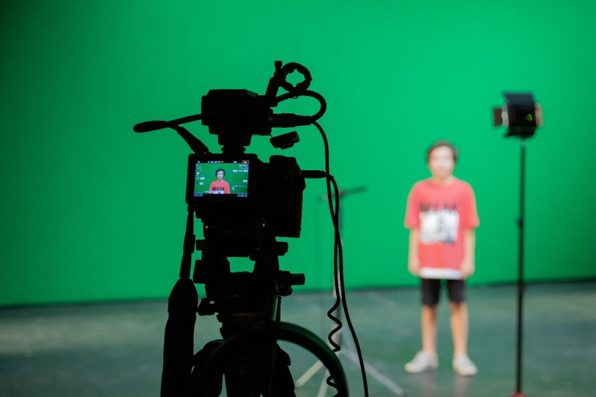 A video camera on a tripod in front of a green screen, with a blurred child standing on the right.