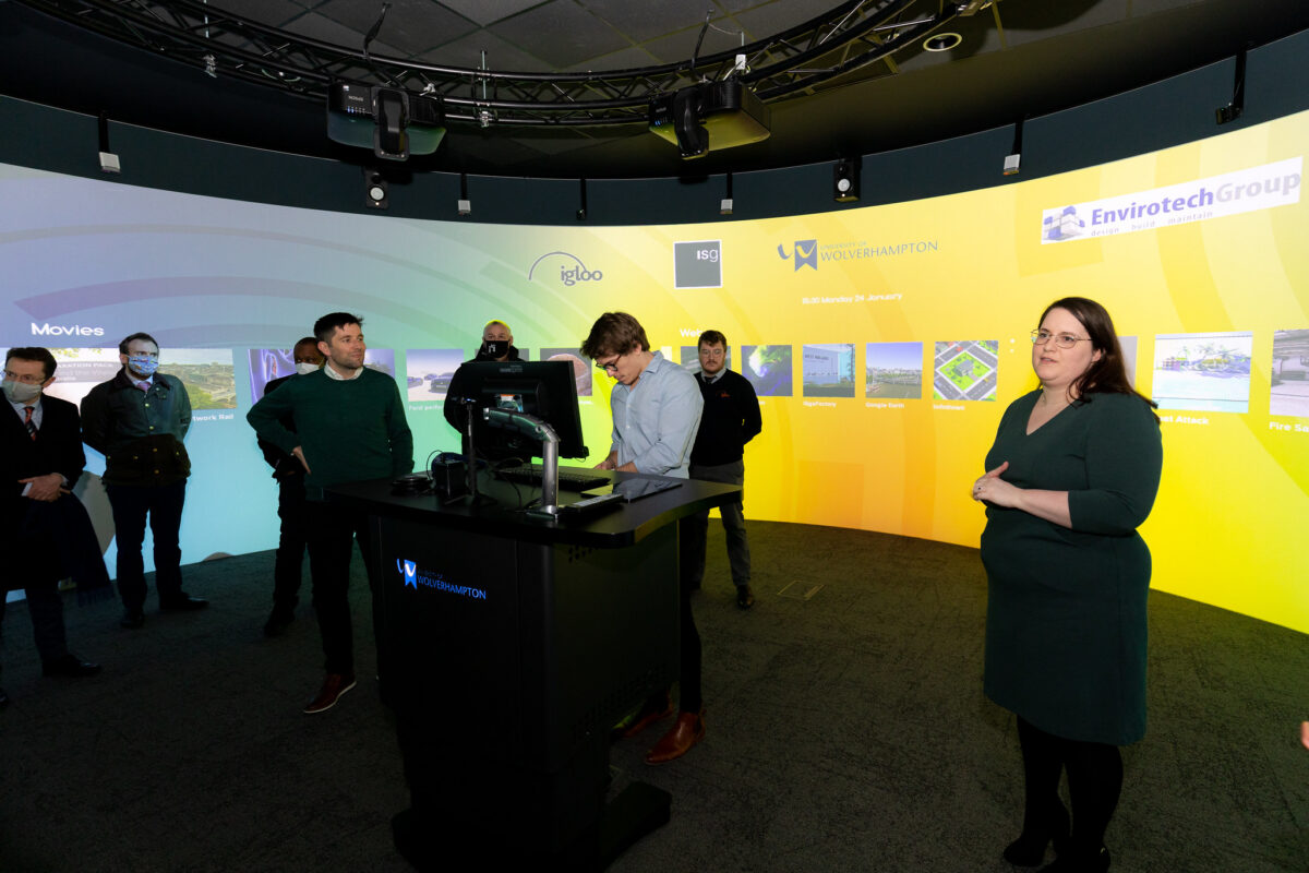 A group of people in a room with a curved screen displaying logos and images, one person is at a computer.