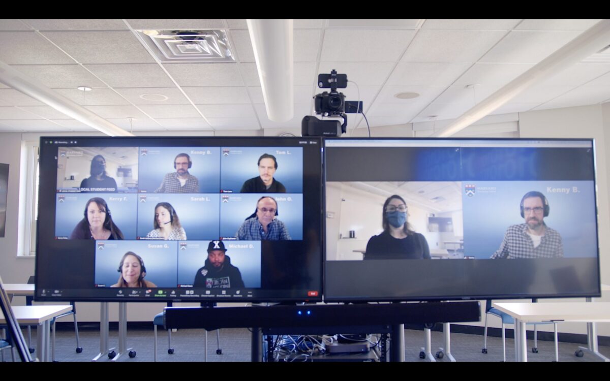 Two large screens showing a video conference with multiple participants, and a camera on a tripod