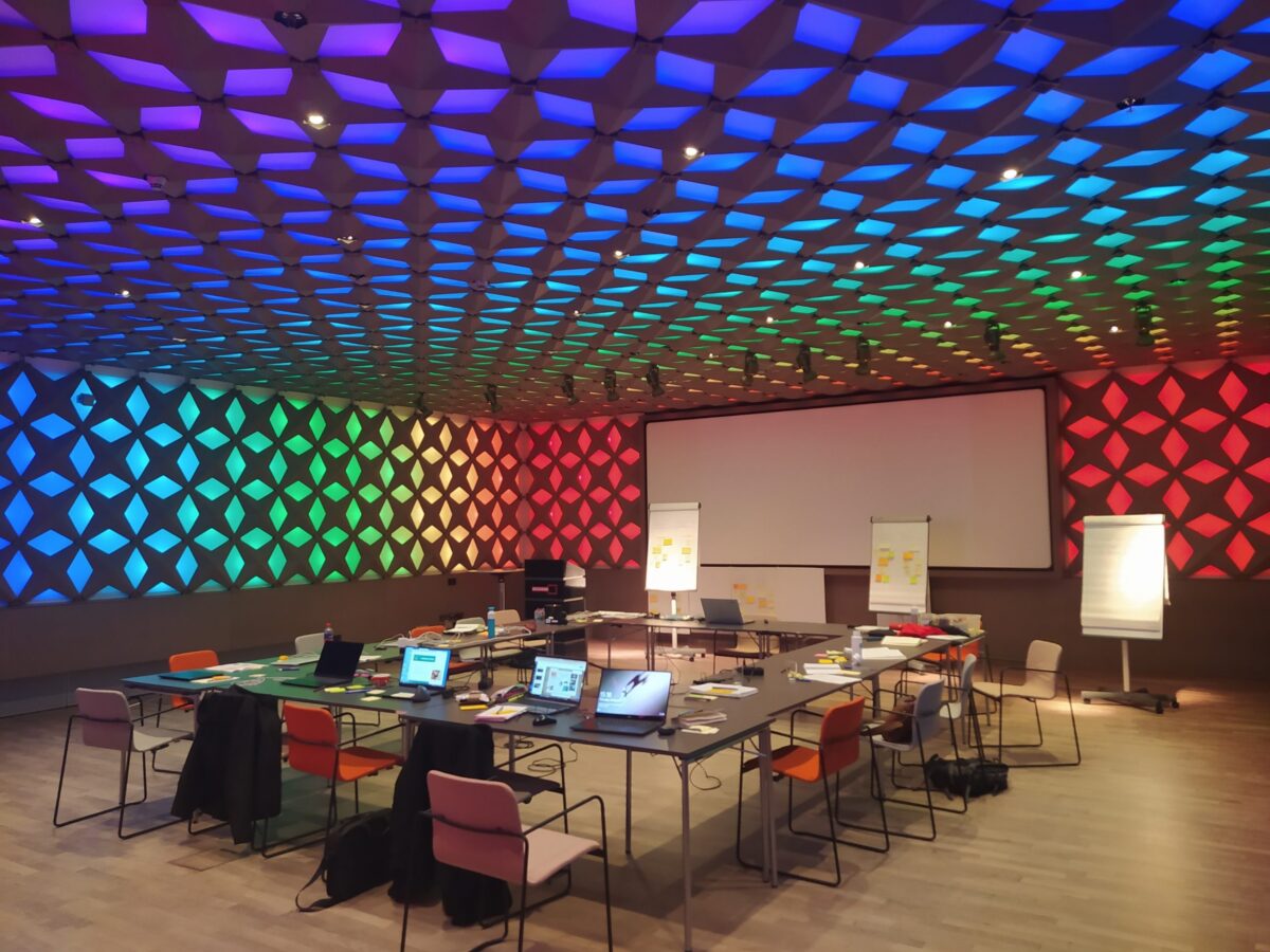 A conference room with a U-shaped table arrangement, laptops, and a large screen, lit by rainbow-coloured wall and ceiling