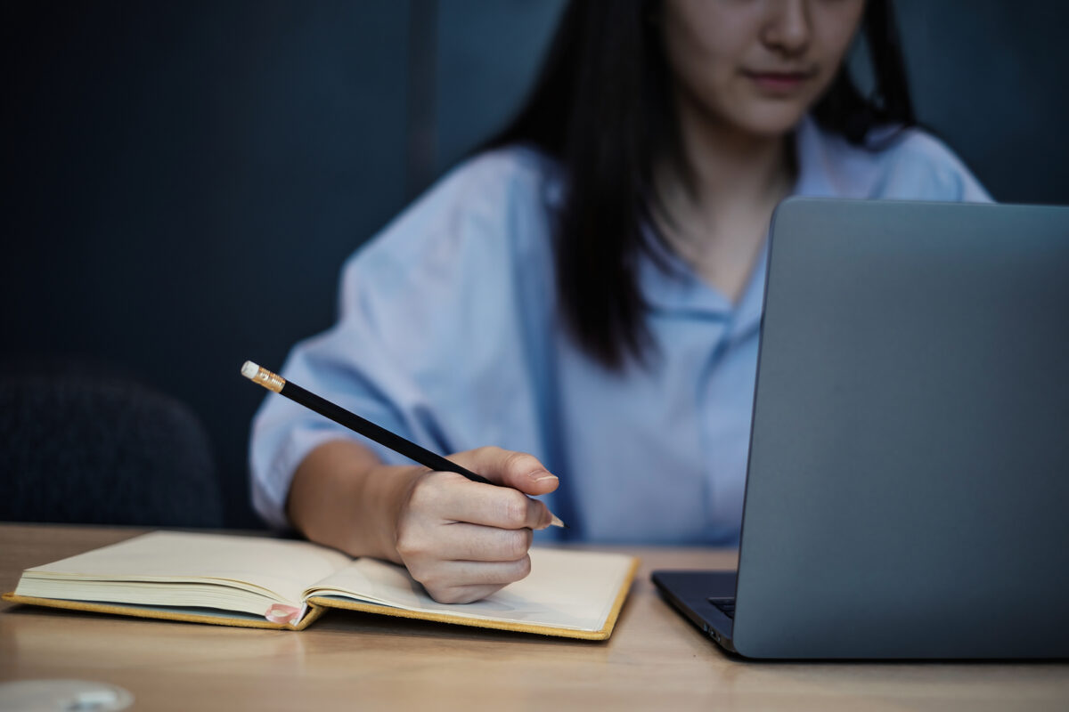 A person in a light blue shirt writing in a notebook with a pencil, next to a laptop.