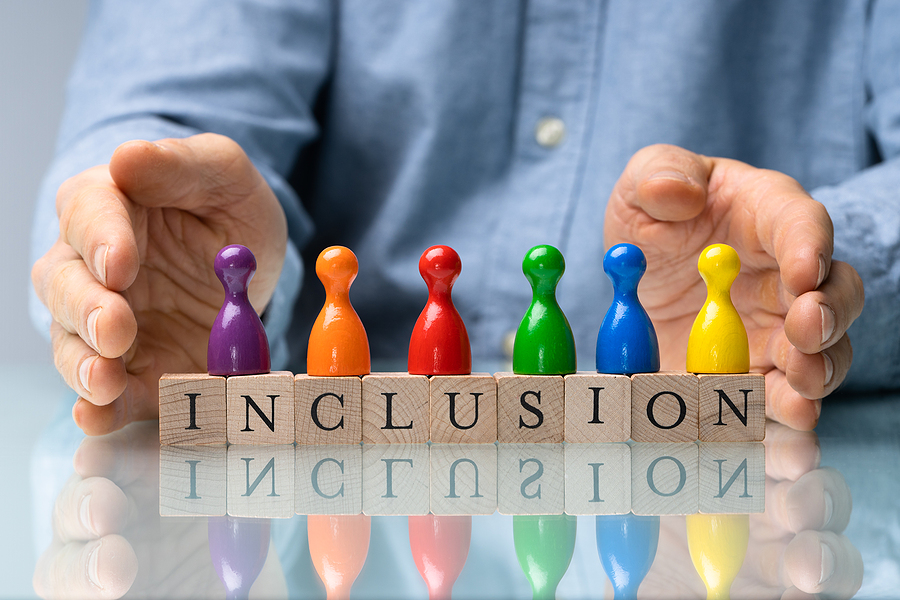 Hands protect six colourful pawns standing on wooden blocks spelling "INCLUSION" on a reflective surface.