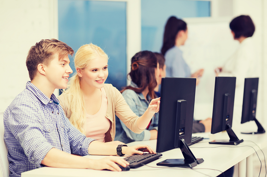 Two students, a blonde woman and a man, smiling and working together on a computer in a classroom.