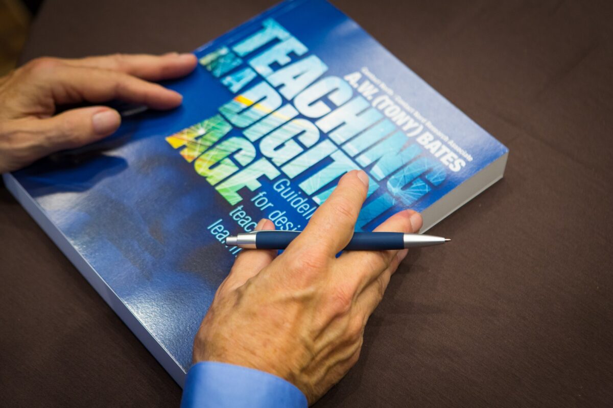 A person's hands holding a book titled 'Teaching in a Digital Age' by A.W. (Tony) Bates, with a pen.