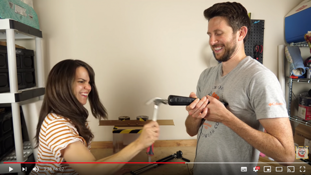 A man and a woman laughing. The woman is holding a hammer, the man is holding a black object.