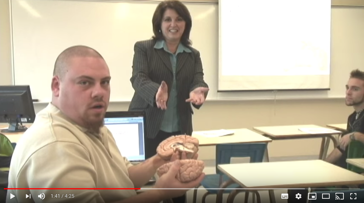 A male student holding a brain model in a classroom, with a female teacher and another student.