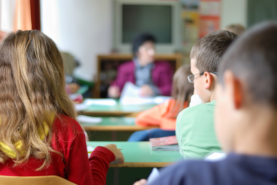 Children in a classroom, seen from behind, with a teacher at the front of the room.