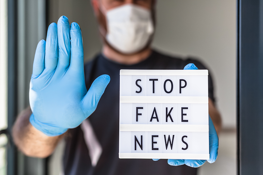 Person wearing a face mask and blue gloves holds up a hand in a stop gesture and a sign saying STOP FAKE NEWS.