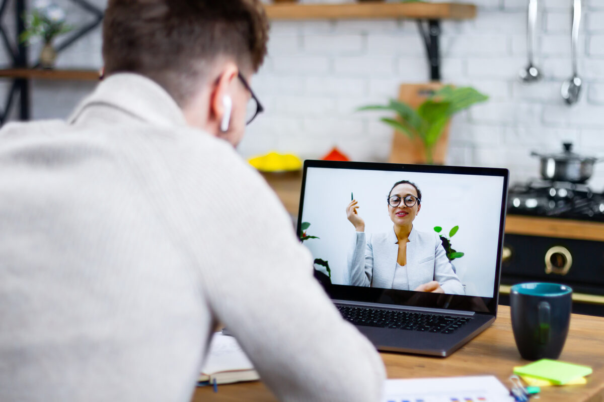 A person with headphones and glasses watching an online lecture on a laptop, a woman on screen.