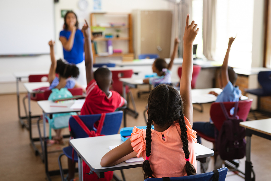 Rear view of students raising hands in a classroom, with a teacher standing at the front.