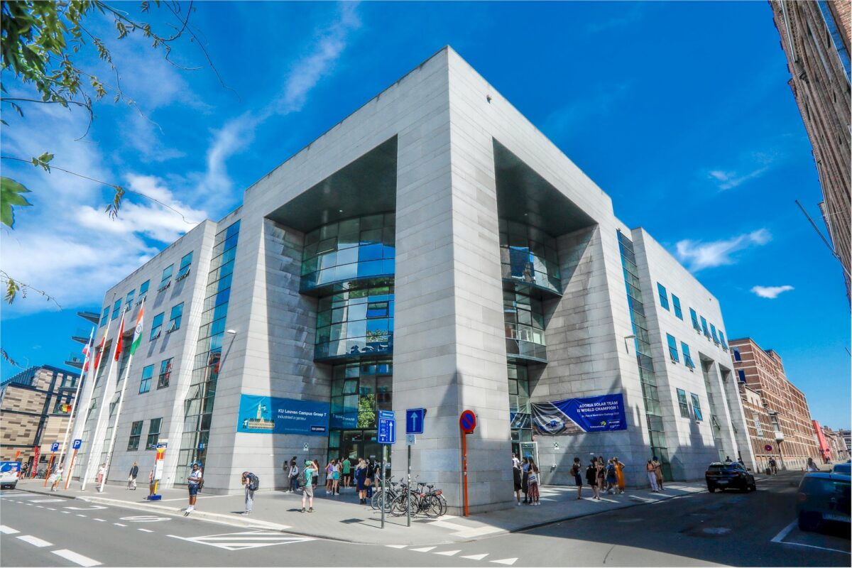 Exterior of a modern building with large glass windows and flags, people gathered outside on a sunny day.