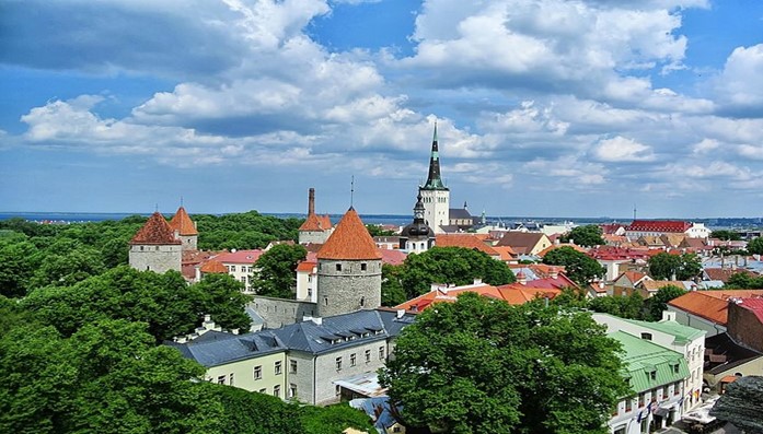 Panoramic view of Tallinn, Estonia, with medieval towers, church spires, and red-tiled roofs amidst green trees.