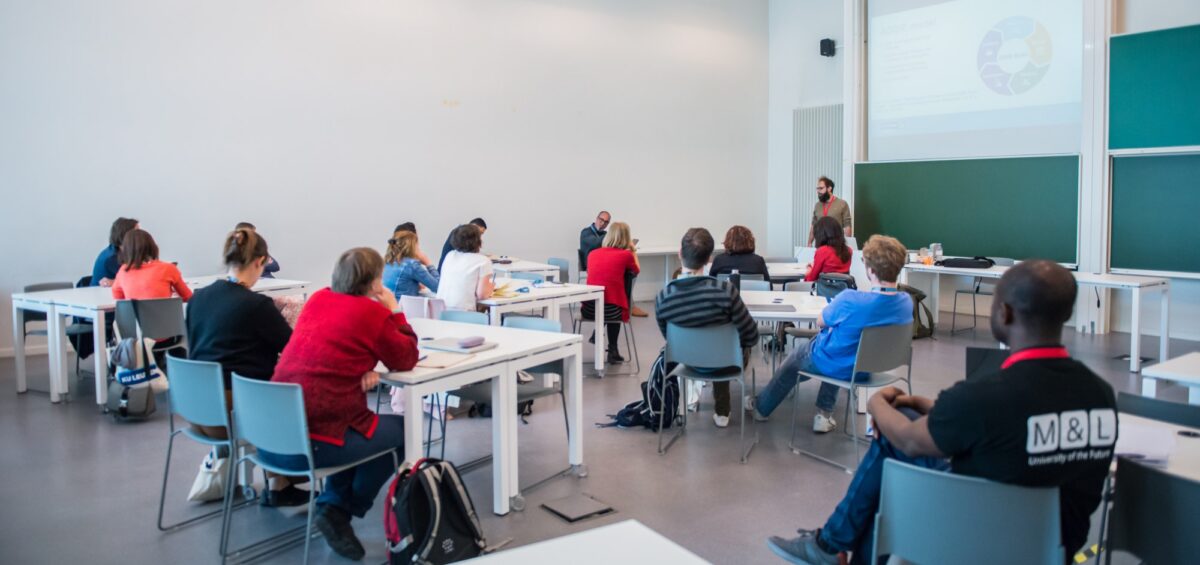 Participants seated at tables in a classroom, facing a speaker and a projected presentation slide.