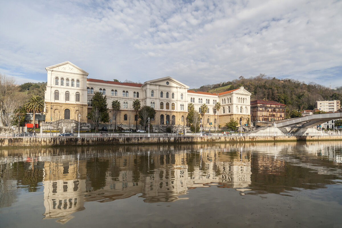 A large, ornate white building with a red roof reflected in a river, with a bridge on the right.