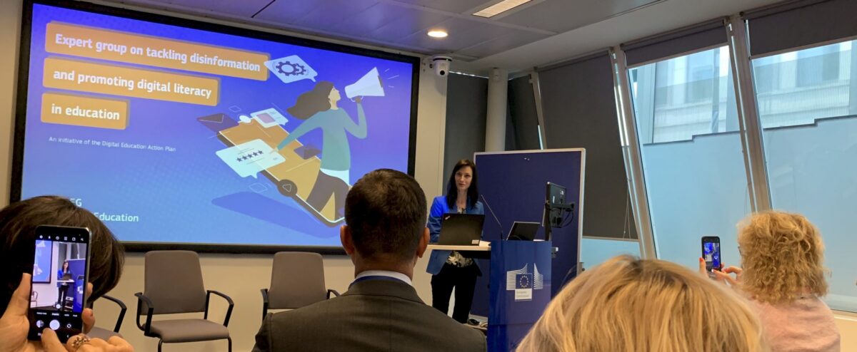 A woman presenting on stage in front of a screen displaying "Expert group on tackling disinformation and promoting digital