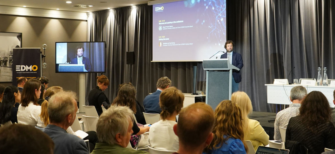 A speaker at a podium presenting to an audience at the EDMO conference, with a large screen showing the agenda.