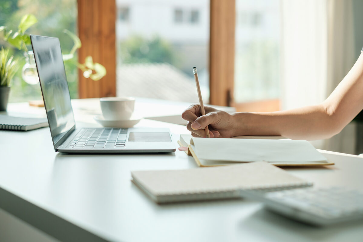 A person's hand writing in a notebook with a pencil, next to a laptop and a cup of coffee.