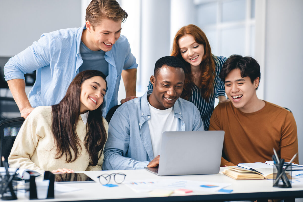 Five smiling students, three men and two women, looking at a laptop screen on a desk.