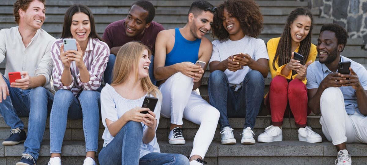 A diverse group of young adults sitting on steps, laughing and looking at their mobile phones.