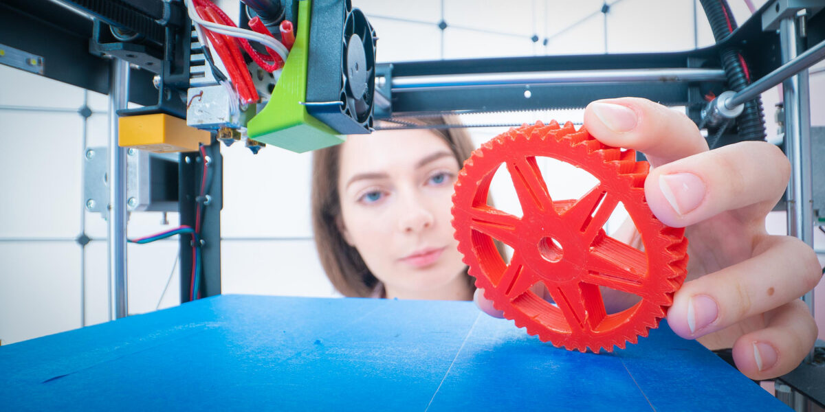 Young woman holding a red 3D-printed gear, with a 3D printer and blue print bed in the foreground.