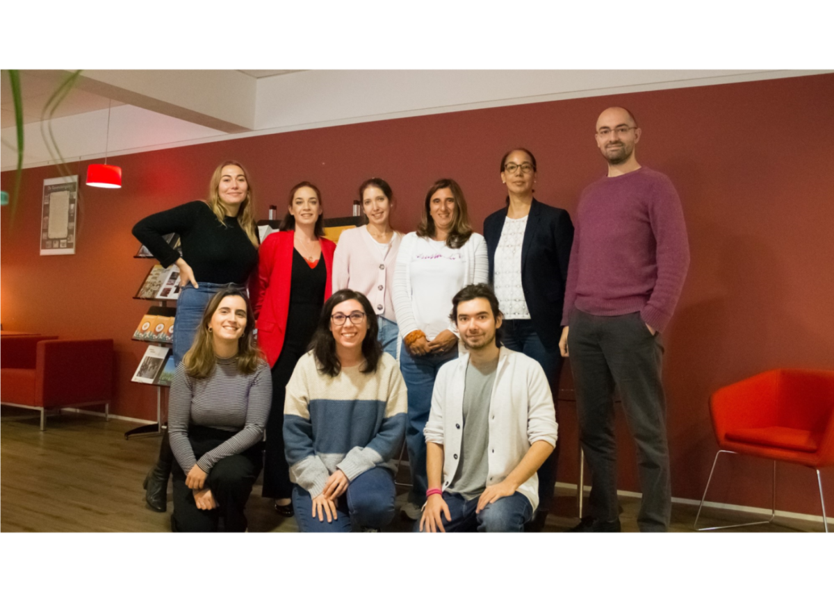 Nine people, seven women and two men, standing and kneeling in a group against a red wall.