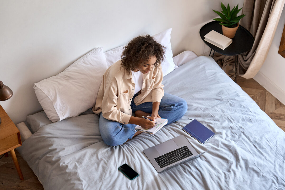 Young woman with curly hair sitting cross-legged on a bed, writing in a notebook. A laptop and phone are next to her.