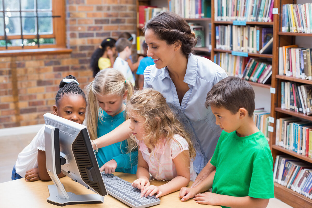 Teacher and four primary school children gathered around a computer in a library, looking at the screen.