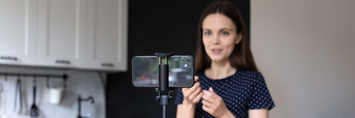 A woman in a blue polka dot top records a video of herself speaking on a smartphone on a tripod.