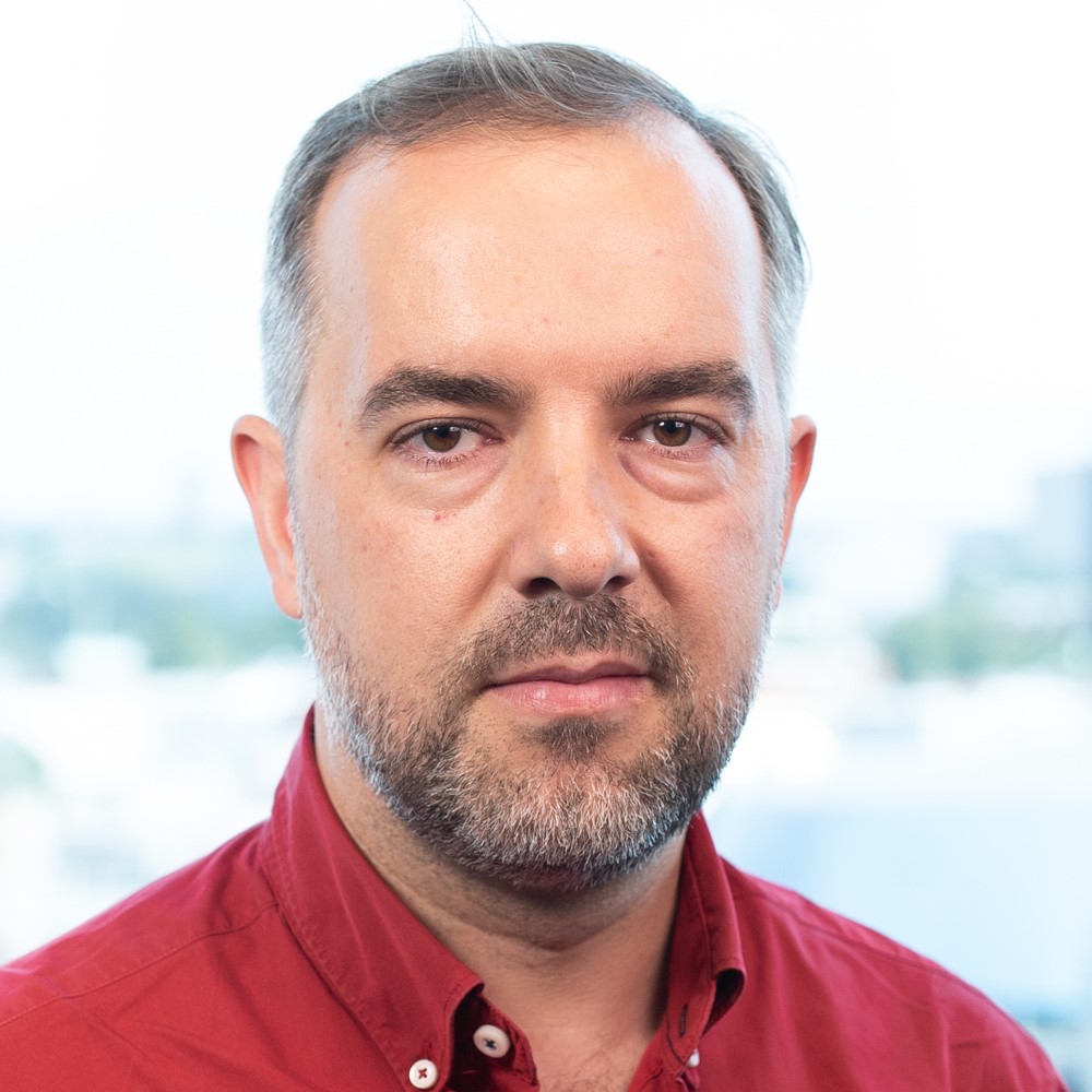 Luis, a man with a beard and short grey hair, wearing a red shirt, looks directly at the camera.
