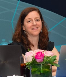 MJB, a woman with brown hair, smiling at the camera, with laptops and a flower arrangement in front of her