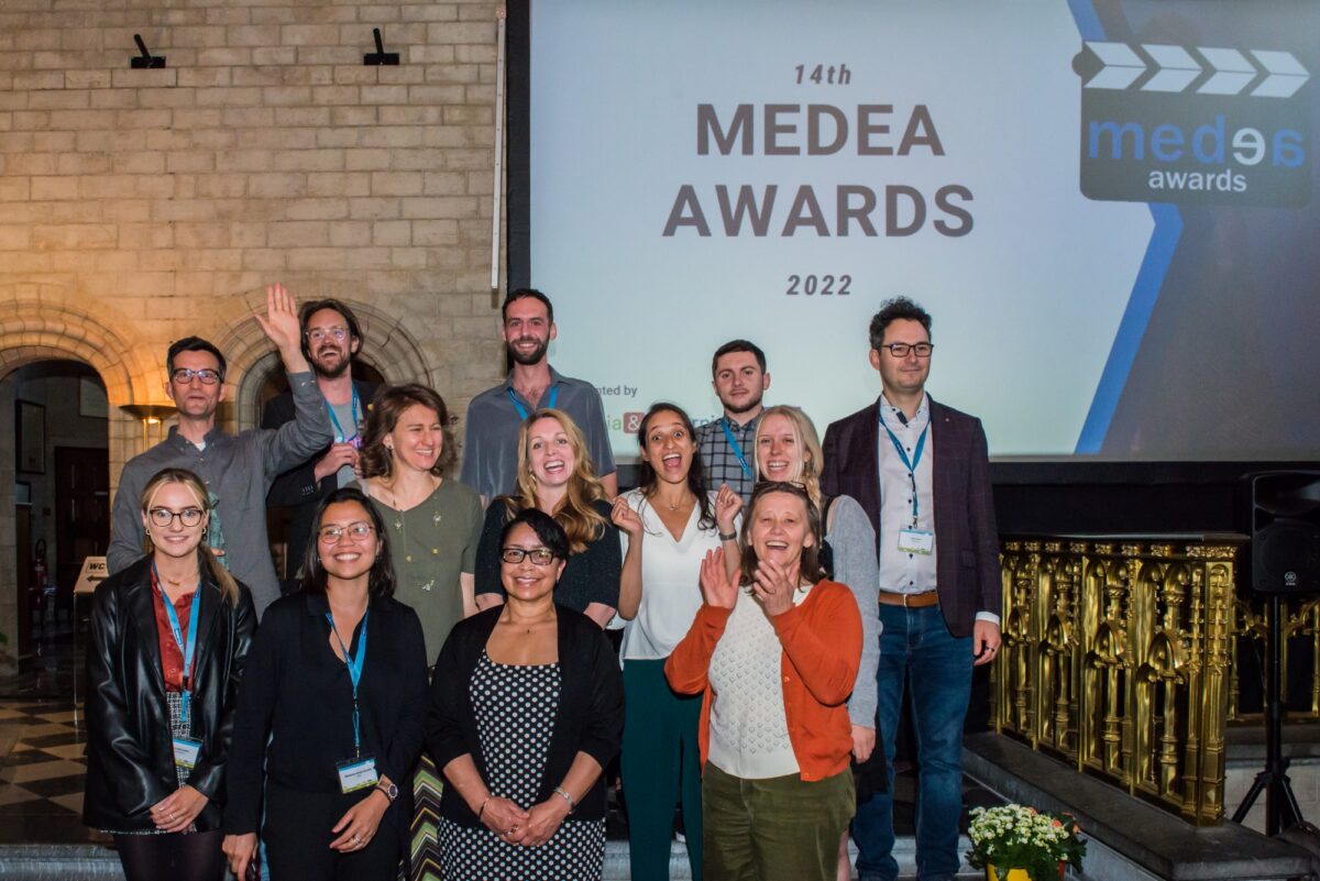 Group photo of 14 people, some cheering, in front of a screen displaying "14th MEDEA AWARDS 2022