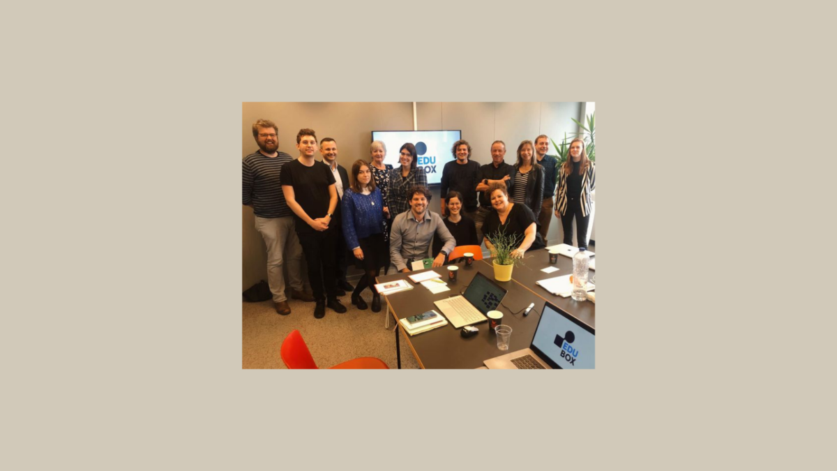A group of people, some standing and some seated around a table, in a meeting room with a screen showing "EDU BOX