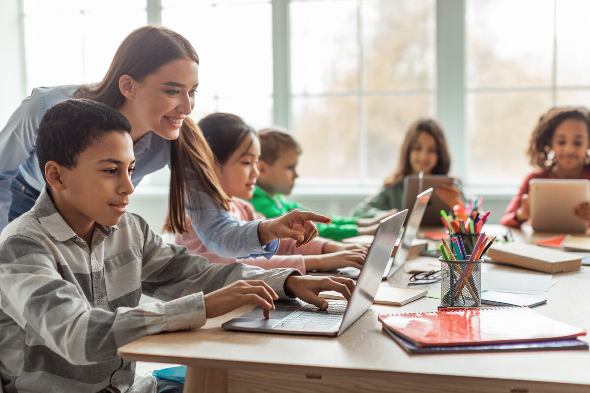 A teacher helps a male student use a laptop in a classroom with other children using devices.